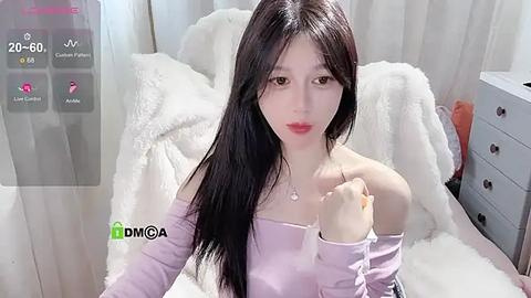 Media: Video of a young East Asian woman with long, straight black hair, fair skin, and red lipstick, wearing a light purple off-shoulder top, sitting on a white fur blanket, with a smartphone overlay displaying a \"DIYA\" watermark.