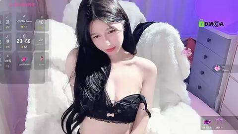 Media: Video of a young Asian woman with long black hair, wearing a black lace bra, sitting on a white fur bedspread in a dimly lit bedroom.