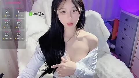 Media: Video of an East Asian woman with long black hair, fair skin, and a slim physique, wearing an off-shoulder white top, sitting on a white fur rug in a dimly lit room.