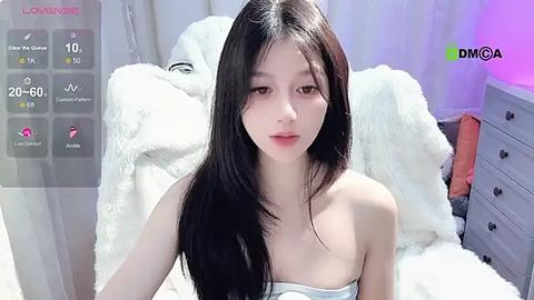 Media: Video of a slender, young Asian woman with long black hair, wearing a strapless dress, sitting on a white bed. The room has a white and purple color scheme, with a fluffy white blanket.