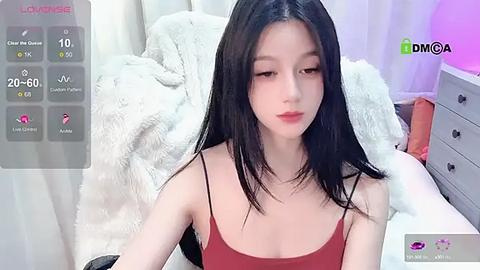 Media: A video of an Asian woman with long black hair, fair skin, and a slender physique, wearing a red spaghetti strap top, sitting on a bed with white fur blankets.