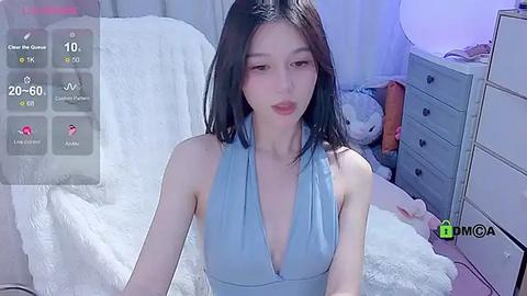 Media: Video of a slim, East Asian woman with long black hair, wearing a blue halter dress, sitting on a white bed with plush toys in a softly lit room.