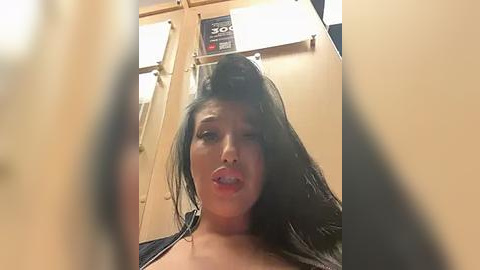 Media: A video of a young woman with long, dark hair, wearing a black top, sticking her tongue out, taken from a low angle. The background shows a beige wall with a white sign and a black trash can.