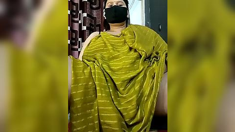 Media: Video of a person wearing a green, patterned saree with a black mask, standing in a room with patterned curtains and a partially visible door.