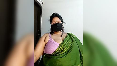 Media: Video of a plus-sized South Asian woman in a green sari, black mask, and pink bra, standing in a doorway, with a blurred person in the foreground.