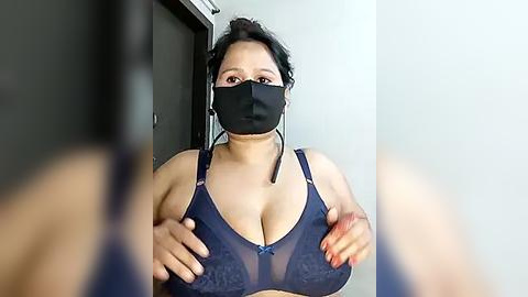 Media: Video of a curvy woman with light skin, black hair, and large breasts, wearing a black mask and a navy blue lace bra, standing in a modern room.