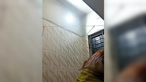 Media: Video of a small, dimly lit room with beige wavy-tile walls, a brown leather couch, and a barred window. The lighting is sparse, creating a somber, claustrophobic atmosphere.