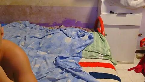 Media: Video of a messy bedroom with a blue and white polka-dot bedspread, a green and white striped pillow, and a red and white striped blanket. The room has a worn-out wall and a white dresser with a red chair.