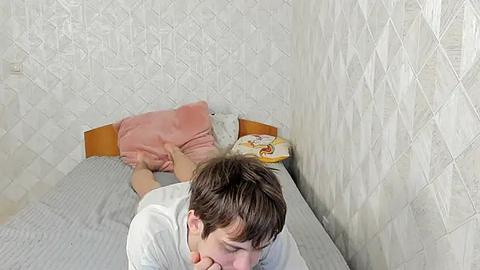 Media: Video of a man in a white shirt, lying on a bed with gray bedding, against a patterned white wall, performing oral sex on a woman in pink underwear.