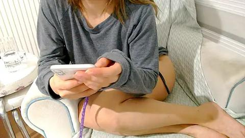 Media: A video shows a young person with fair skin, wearing a gray sweatshirt and purple underwear, sitting on a white chair, engrossed in a white smartphone.