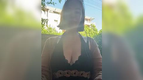 Media: A video of a woman with a short bob haircut, wearing a black lace bra under a sheer beige cardigan, standing in a garden with greenery and a house in the background.