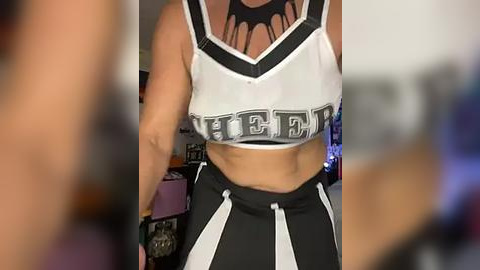 Media: Video of a woman in a white and black striped sports bra with \"3\" on the chest and black shorts, showing a toned midriff. Background is blurred with shelves and colorful objects.