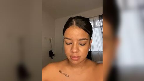 Media: Video of a young, light-skinned woman with dark hair, wearing makeup, with a tattoo on her collarbone. She is topless, in a dimly lit room with white walls and a window.