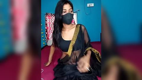 Media: Video of a woman with long black hair, wearing a black mask, black saree, and gold shoulder strap, sitting on a pink bed, in a blue room with a patterned cushion.