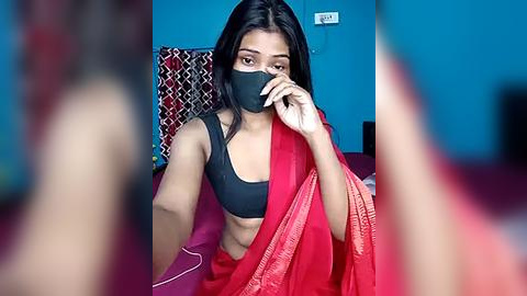 Media: Video of a South Asian woman with long black hair, wearing a black bra and red sari, black face mask, seated on a bed with a red pillow, in a blue room with a white light switch.