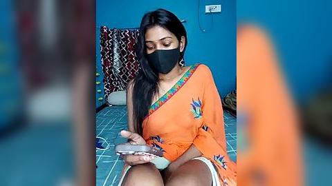 Media: Video of a young South Asian woman with medium brown skin, long black hair, wearing an orange sari with a colorful border, black mask, and seated on a bed in a blue-painted room.