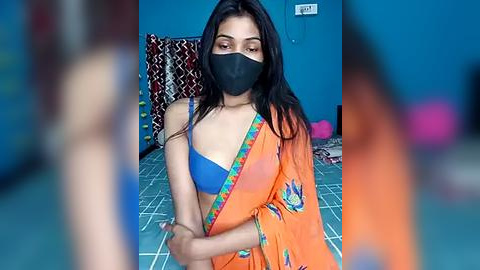 Media: A video of a young South Asian woman with long black hair, wearing an orange sari, blue bra, black face mask, and a blue t-shirt, in a room with a tiled floor, patterned curtains, and a blue wall.