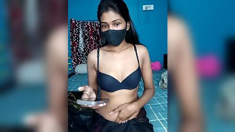 Media: Video of a young, slender South Asian woman with long black hair, wearing a black bra, mask, and black pants. She sits on a bed with patterned sheets, holding a phone, in a room with blue walls.