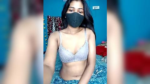 Media: Video of a young woman with medium skin tone, wearing a black face mask, gray lace bra, and blue patterned shorts, sitting on a bed with colorful pillows in a blue-walled room.