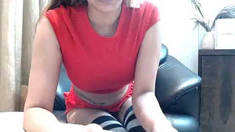 Media: Video of a light-skinned woman with long brown hair, wearing a red crop top and matching shorts, sitting on a black leather couch in a room with beige curtains and a potted plant.