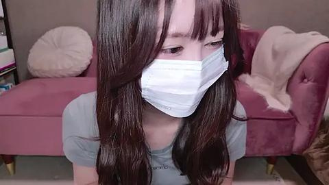 Media: Video of a young Asian woman with long, straight brown hair, wearing a light blue t-shirt and a white surgical mask, sitting indoors on a pink couch with white throw pillows and a beige blanket.