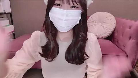 Media: Video of a young East Asian woman with long dark hair, wearing a white surgical mask, white top, and cat ears, seated on a pink velvet sofa.