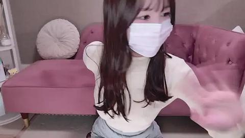 Media: Video of an Asian woman with long black hair, wearing a white face mask, gray pants, and a white sleeveless top, standing in front of a pink tufted couch, indoors.