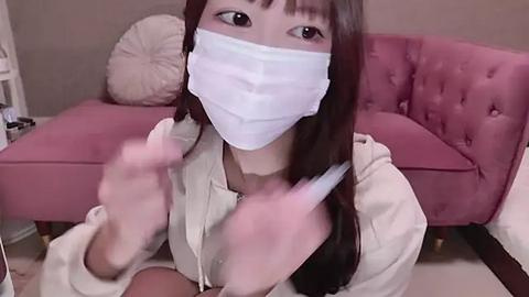 Media: Video of an East Asian woman with long brown hair, wearing a white face mask, beige hoodie, and white gloves, kneeling in a pink velvet armchair, indoors.