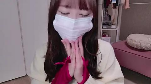 Media: A video of an Asian woman with long brown hair, wearing a white surgical mask, praying with hands clasped in a room with a pink couch and open closet filled with clothes.