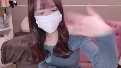 Media: Video of an Asian woman with long brown hair, wearing a white mask, blue sweater, and jeans, sitting on a bed, blurred background.