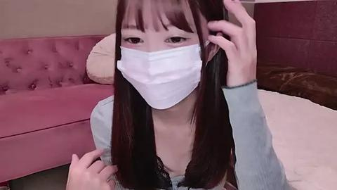 Media: Video of an East Asian woman with long black hair, wearing a white surgical mask and light blue top, sitting on a pink couch with a beige pillow.