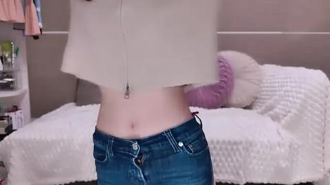 Media: Video of a person, likely female, holding a white towel, wearing blue jeans, standing in a bedroom with a white bed, purple pillow, and a closet visible in the background.