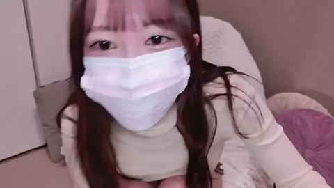 Media: A video of an Asian woman with long, dark hair and pale skin, wearing a white mask, sitting on a bed with light-colored sheets, looking directly at the camera.