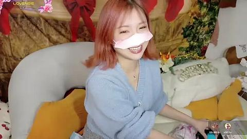 Media: A young Asian woman with light pink hair, wearing a blue cardigan and a pink face mask, smiles while sitting on a yellow cushioned bench. The background features festive decorations and a plush toy.
