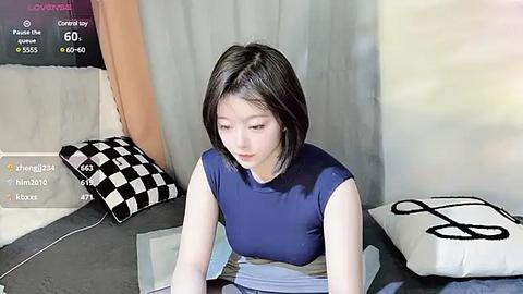 Media: A video of a young Asian woman with straight black hair, wearing a blue sleeveless top, sitting in a virtual reality headset in a room with gray and white walls, checkered pillows, and a black and white cushion.