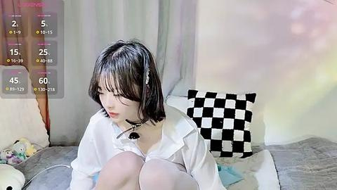 Media: A video shows an East Asian woman with short black hair, wearing a white shirt and black skirt, seated on a grey sofa. The room features a checkered cushion and a stuffed animal in the background.