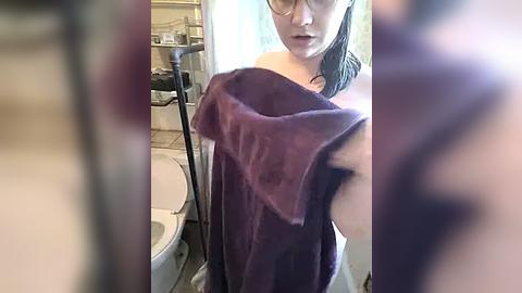 Media: A video shows a woman with glasses, wet hair, holding a purple towel, standing in a bathroom with a toilet and sink in the background.