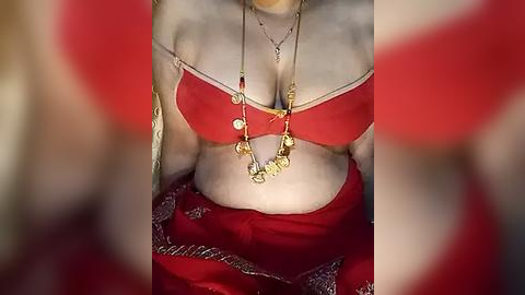 Media: Video of a woman in a red bikini top with gold necklaces, seated, blurred background.