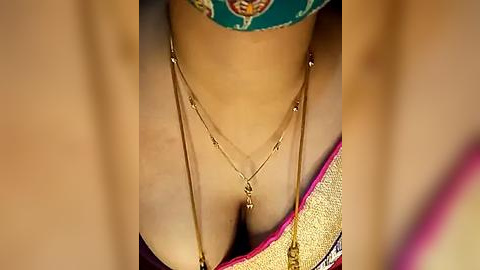 Media: Close-up video of a woman's neck and cleavage, adorned with two gold necklaces featuring small pendants. She wears a colorful, patterned headscarf and a gold necklace with a pendant.