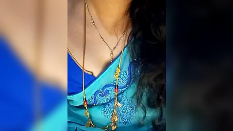 Media: A close-up video of a woman wearing a vibrant turquoise blouse with intricate blue embroidery and a gold necklace with red beads, blurred background.