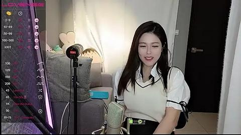 Media: Video of an East Asian woman with long black hair, wearing a white sweater and black skirt, singing into a microphone in a cozy living room with a gray couch and a lamp.