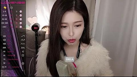 Media: Video of a young East Asian woman with straight, dark hair, wearing a fur coat, sitting in a cozy room with soft lighting and a microphone, engaged in an intimate conversation.