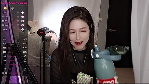 Media: Video of an East Asian woman with long black hair, wearing headphones, singing into a blue microphone. Background features a plush toy and recording equipment, in a dimly lit room.