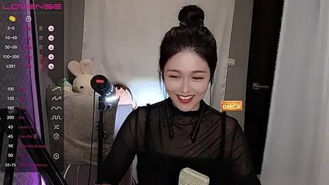 Media: Video of an East Asian woman with fair skin, dark hair in a bun, wearing a sheer black top, smiling while holding a microphone. Background features plush toys and a digital clock, with a \"LOUNGE\" banner.