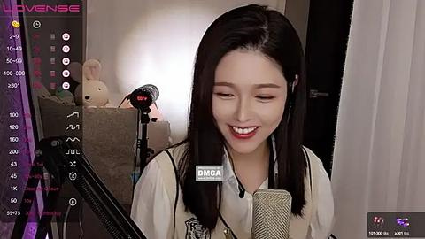 Media: A video of an East Asian woman with straight black hair and fair skin, smiling while singing into a microphone, surrounded by a dimly lit room with a plush teddy bear and studio equipment.