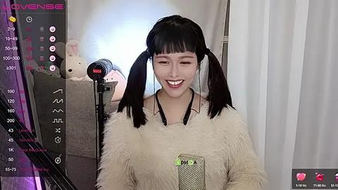 Media: Video of a smiling East Asian woman with pigtails, wearing a fuzzy beige sweater, singing into a microphone in a cozy, dimly lit room with white curtains.