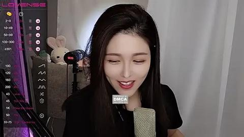 Media: A video of a young Asian woman with long black hair, wearing a black shirt, singing into a microphone. She has fair skin, a gentle smile, and is surrounded by soft lighting and plush toys.