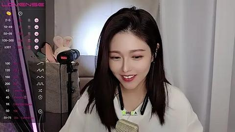 Media: Video of an East Asian woman with straight, shoulder-length dark hair, wearing a white robe, sitting at a desk, using a microphone and headphones, in a modern, dimly lit room with a plush teddy bear.