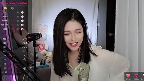 Media: Video of an Asian woman with straight black hair, wearing a white blouse, singing into a microphone in a cozy room with a stuffed bunny and a calendar.