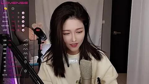 Media: Video of a young Asian woman with long black hair, wearing a white t-shirt, singing into a microphone. Background includes a dark room with a video camera and studio lights.